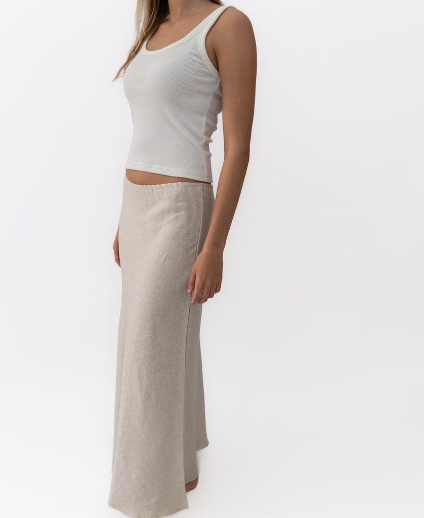 A woman wears the Lazy Beach Pearl Maxi Skirt Linen in natural linen with a white tank top, standing against a plain white background.