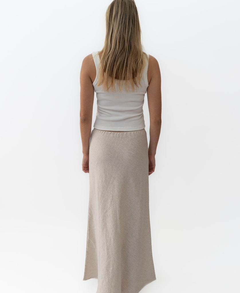 A woman with long blonde hair, wearing a white sleeveless top and the Lazy Beach Pearl Maxi Skirt Linen in Natural Linen, stands facing away against a plain white background.