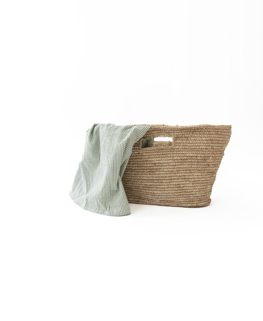 The Lazy Beach Raffia Basket | Natural features woven raffia with rectangular handles, resting on a white surface and partially covered by a light green and white striped cloth—ideal for carrying your beach essentials.