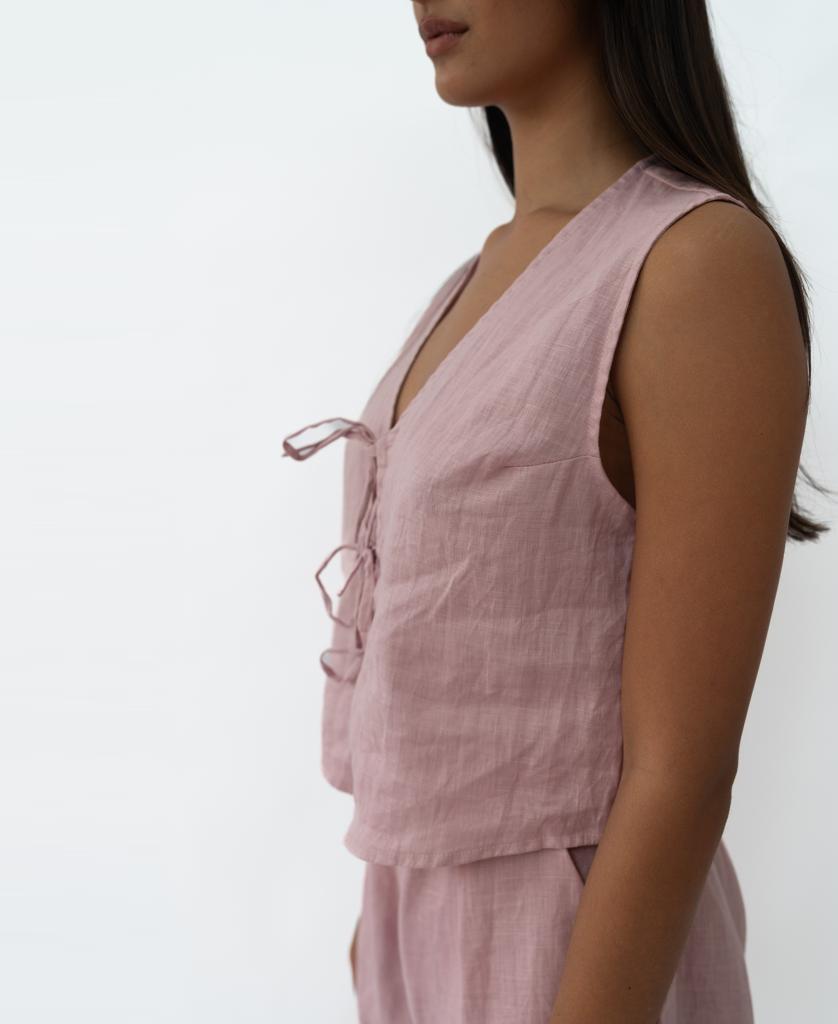 A woman is seen in profile wearing the Lazy Beach Rennie Vest in blush linen with front ties and matching bottoms, capturing effortless summer style against a plain white backdrop.