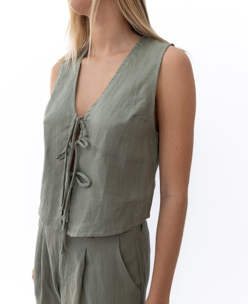 A woman wears the Lazy Beach Rennie Tie Vest Linen in Dark Khaki, a sleeveless V-neck with front ties, handmade in Bali, paired with matching high-waisted pants and shown from the torso up against a white background.