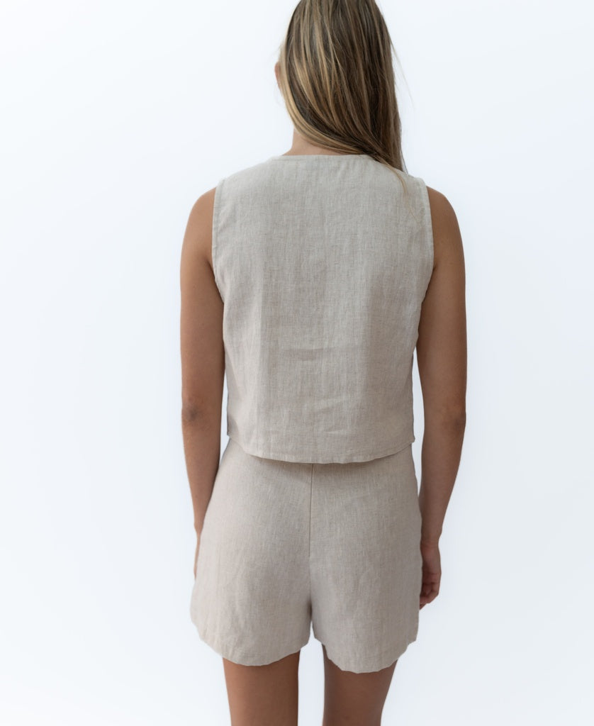A woman with long, straight hair stands with her back to the camera in the Lazy Beach Rennie Tie Vest Linen | Natural against a white background, showcasing this timeless, comfortable Bali-inspired style.