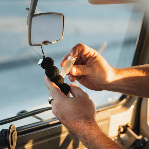 A person uses Smelly Balls Onyx Set Cut Throat, applying fragrance oil from a small bottle onto ethically sourced wool felt balls that hang from a car’s rearview mirror.