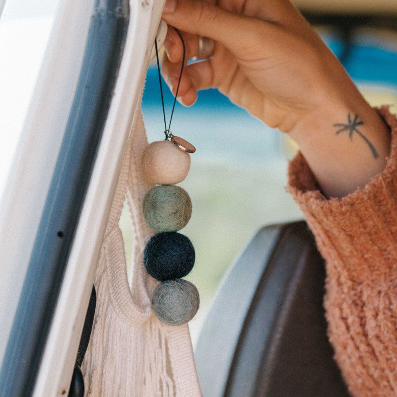 A hand with a star tattoo hangs the Smelly Balls Cove Set Coastal Drift—four wool felt balls in varying shades—from a car door handle, perfect for pairing with your favorite fragrance oil.