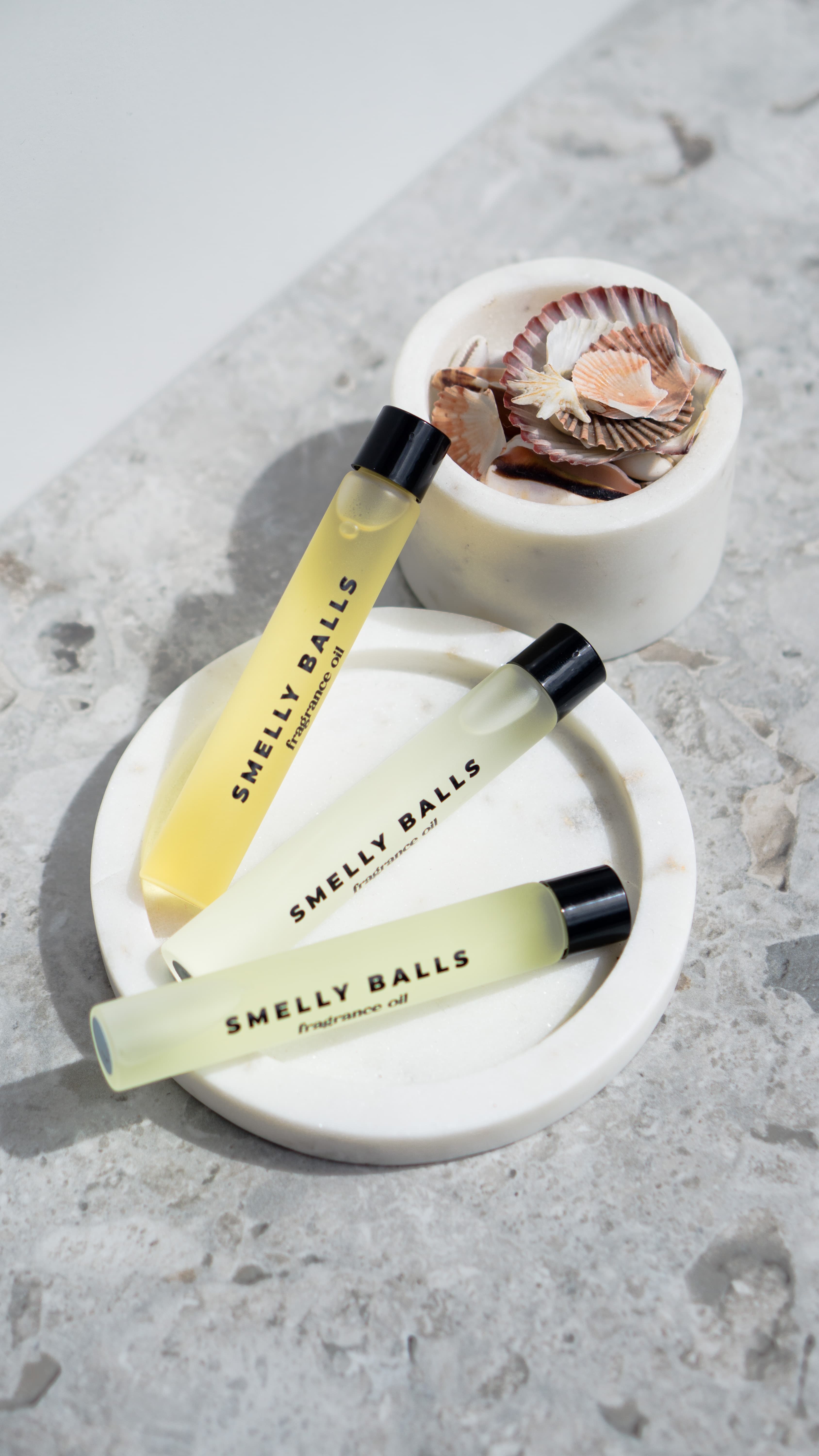 Three tubes of Smelly Balls Sweet Pea Fragrance Oil are arranged on white circular trays with seashells, set atop a gray textured surface.