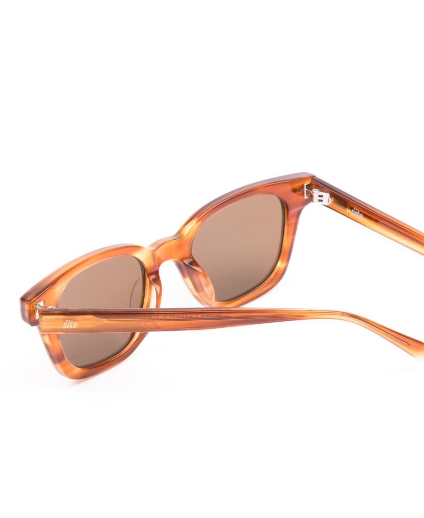 The Sito Ryker sunglasses in Tangelo/Brown Polar feature a plant-based acetate frame and dark lenses, shown angled on a white background to display the arms. Enjoy UV protection with sustainable style.