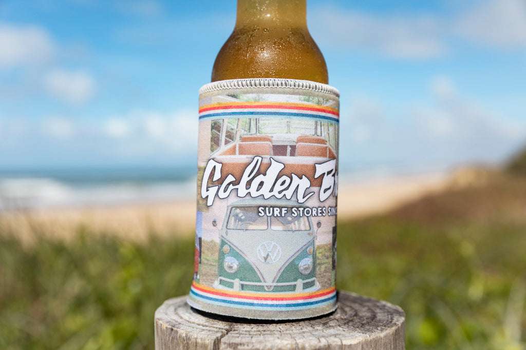 A GB Kombi Collage Stubby by Golden Breed, featuring a colorful Kombi design, holds a beer bottle on a wooden post by the beach.