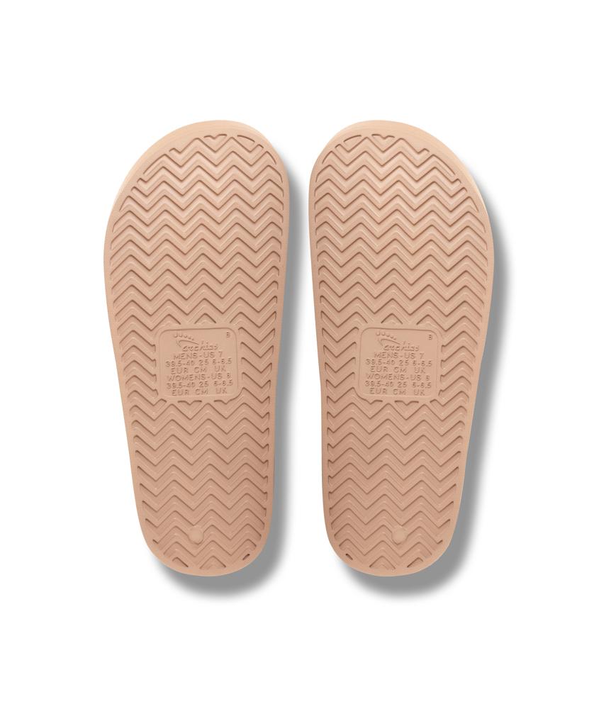 Two Archies Archie Slide | Tan soles made from closed cell foam feature a herringbone tread and embossed rectangular text in the center, shown side by side on a white background.