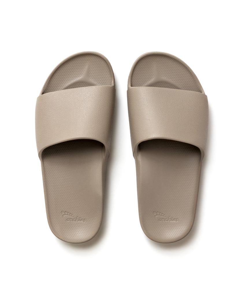 Archies Archie Slide in Taupe: A pair of beige slide sandals with wide straps, contoured footbeds for comfort, and open toes, shown from above on a white background.