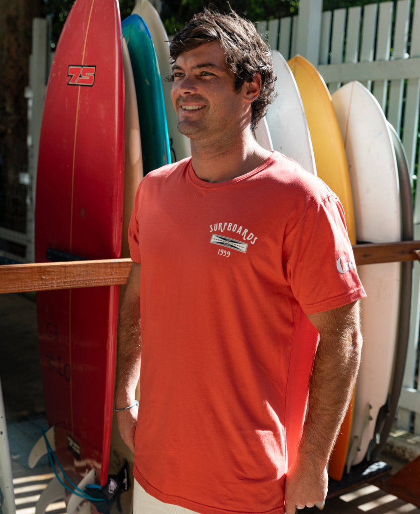 A man in a wash red G&S Surfboards Tee by Gordon & Smith stands smiling in front of several upright surfboards on a sunny day.