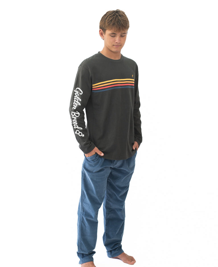 A young man wearing a Heavy Island Long Sleeve Tee | Wash Black with horizontal yellow, orange, and blue stripes across the chest and "Golden Breed" text on the sleeve, paired with blue pants, standing barefoot against a white background.