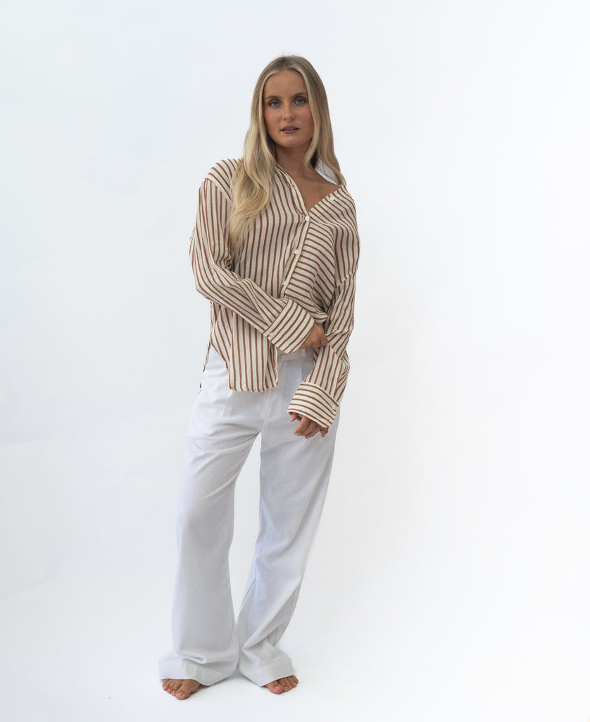 Woman wearing a relaxed fit Miley | Chestnut striped shirt paired with loose white pants, standing barefoot against a plain light background in a casual, retro style.