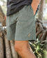 A person in a dark t-shirt wears Golden Breeds Floyd Elastic Waist Walkshort in Khaki, standing outdoors with one hand in the pocket near tree roots.