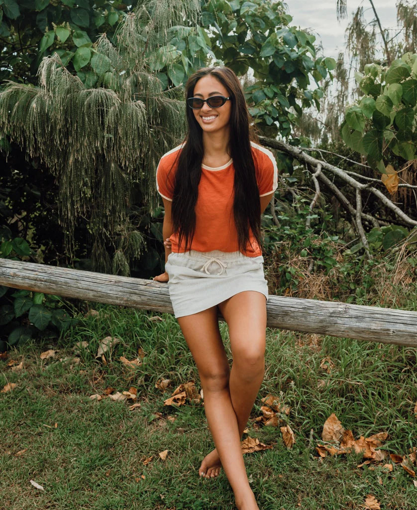 Woman wearing Retro Ringer Tee | Wash Red Beige with sunglasses, sitting outdoors on a wooden rail surrounded by green foliage and trees.
