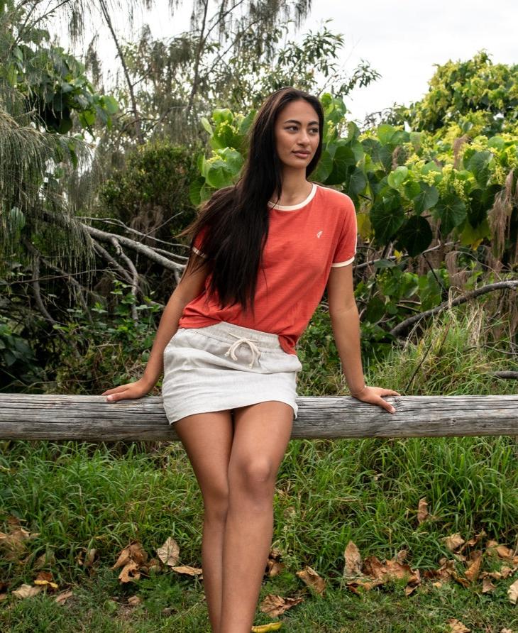 Woman wearing a Retro Ringer Tee | Wash Red Beige with cream trim, sitting on a wooden fence in a lush green outdoor setting, embodying casual Australian surfwear style.