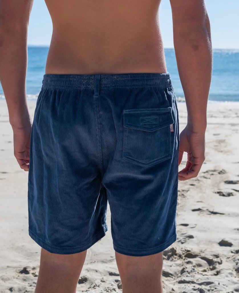 Rear view of a person wearing Floyd Cord Elastic Waist Walkshort | Vintage Navy standing on a sandy beach with the ocean in the background, showcasing the cord fabric and elastic waistband.