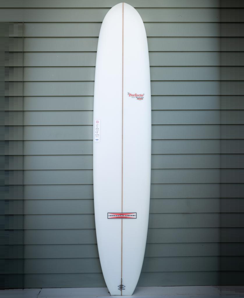 The Gordon & Smith G&S 96 Perfecto Polish Finish longboard in white, featuring a tan stringer and ideal for nose riding, stands upright against a gray paneled wall.