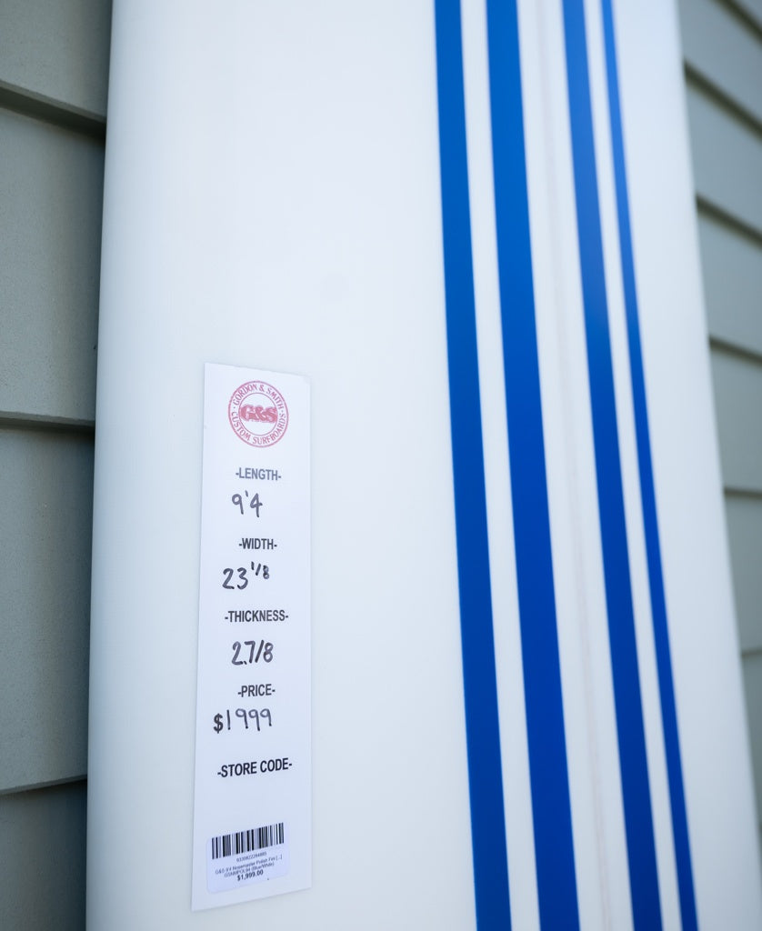 Close-up of the Gordon & Smith G&S 94 Nosemaster Polish Finish longboard in Blue/White, priced at $1999. Ideal for nose riding, it leans against a gray wall, showcasing its sleek design and dimensions.