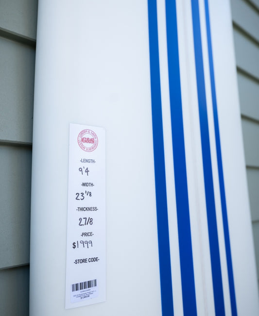 Close-up of the Gordon & Smith G&S 94 Nosemaster Polish Finish longboard in Blue/White, priced at $1999. Ideal for nose riding, it leans against a gray wall, showcasing its sleek design and dimensions.