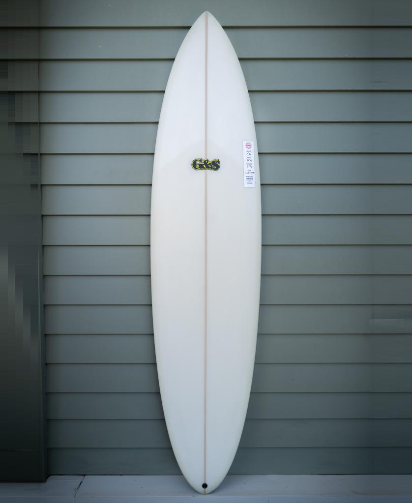A G&S 76 Cool Tool Mid Twin surfboard by Gordon & Smith in off white, celebrated for its wave-catching prowess, stands upright against a gray horizontal siding wall.