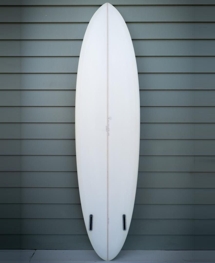 The G&S 76 Cool Tool Mid Twin by Gordon & Smith, in off white with two black fin boxes, stands upright against a green horizontal panel wall, renowned for its excellent wave catching performance.