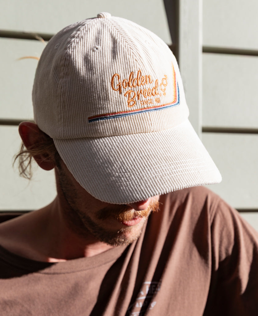Someone wearing the Golden Breed Hancock Cord Cap in Off White sits in sunlight, the brim of the corduroy cap casting partial shade over their face.