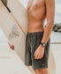 A shirtless person wearing Golden Breeds Floyd Elastic Waist Walkshort in Wash Black and a wristwatch holds a surfboard on a sandy beach, with the ocean and rocks in the background.