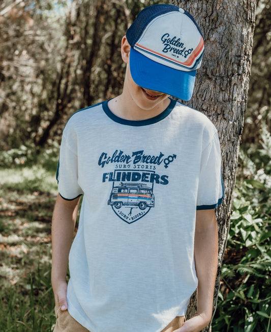 A child wearing a Flinders Rider Kids Tee in Off White by Golden Breed and a cap stands outdoors by a tree, looking down with hands in pockets.