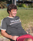 A man with brown hair wearing a Golden Breed Retro Ringer Tee in Wash Black and red shorts sits on a low stone wall outdoors in sunlight, giving off strong 70s vibes.