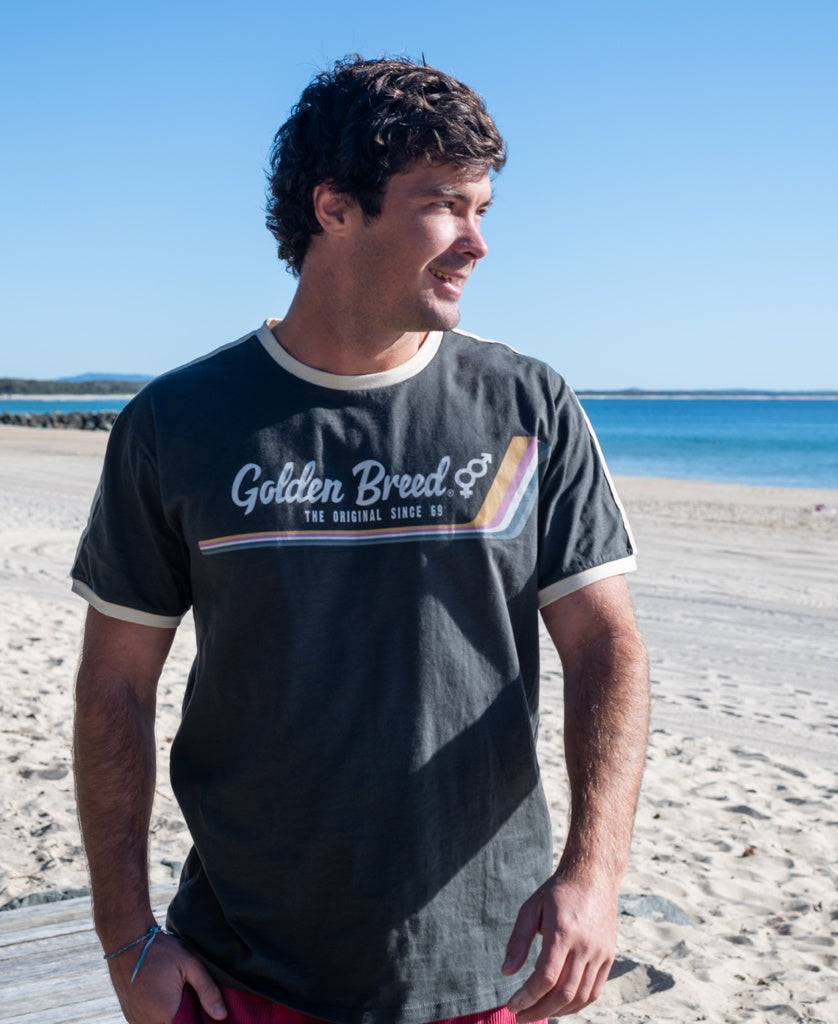 A man on a sandy beach looks left under a clear blue sky, wearing the Golden Breed Retro Ringer Tee in Wash Black and channeling classic 70s style.