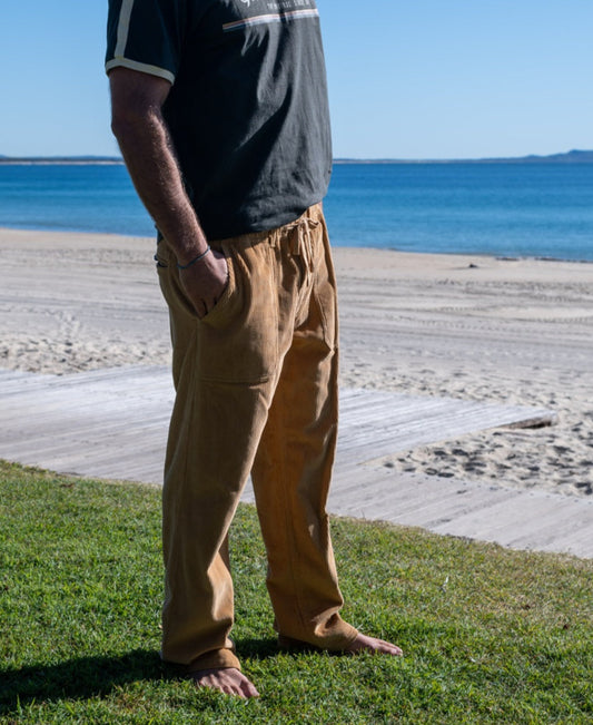 A person in a black t-shirt and Golden Breed George Cord Pant in Dark Camel stands barefoot on grass near a sandy beach with the ocean in the background.