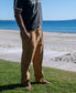 A person in a black t-shirt and Golden Breed George Cord Pant in Dark Camel stands barefoot on grass near a sandy beach with the ocean in the background.