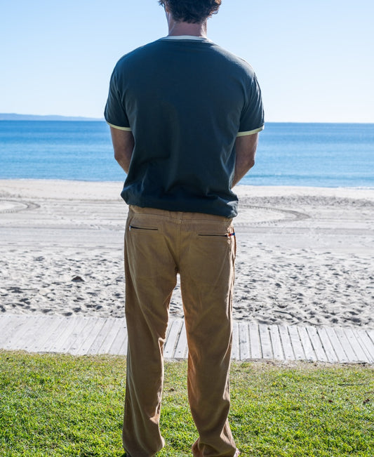 A person in a dark T-shirt and Golden Breed George Cord Pant | Dark Camel, made from 100% cotton, stands on grass facing the calm sea and clear sky.
