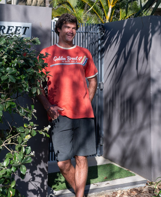 A man wearing the Golden Breed Retro Ringer Tee in Wash Red and dark shorts stands by a gray gate, sunlight casting partial shade, lush greenery around him—channeling classic 70s vibes.