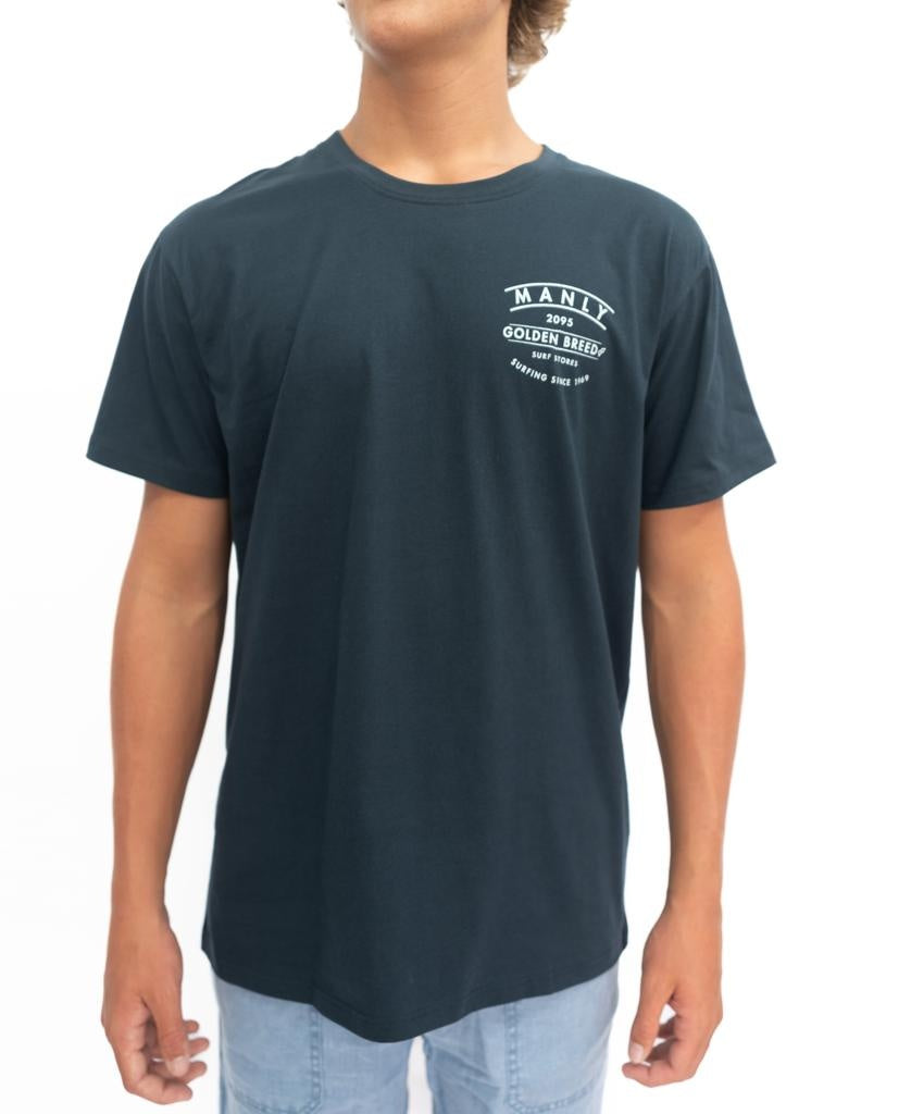 A person wearing a navy Manly Boards Tee by Golden Breed, featuring a white “Manly Boys Golden Birds” logo on the left chest, paired with light blue jeans, stands against a plain white background.