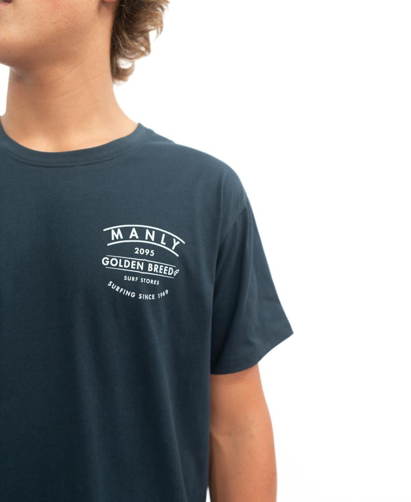 Someone wearing the Golden Breed Manly Boards Tee in navy, featuring bold white Manly 2095 Golden Breed Surf Stores Surfing Since 1969 text—ideal for capturing a classic surf town vibe.