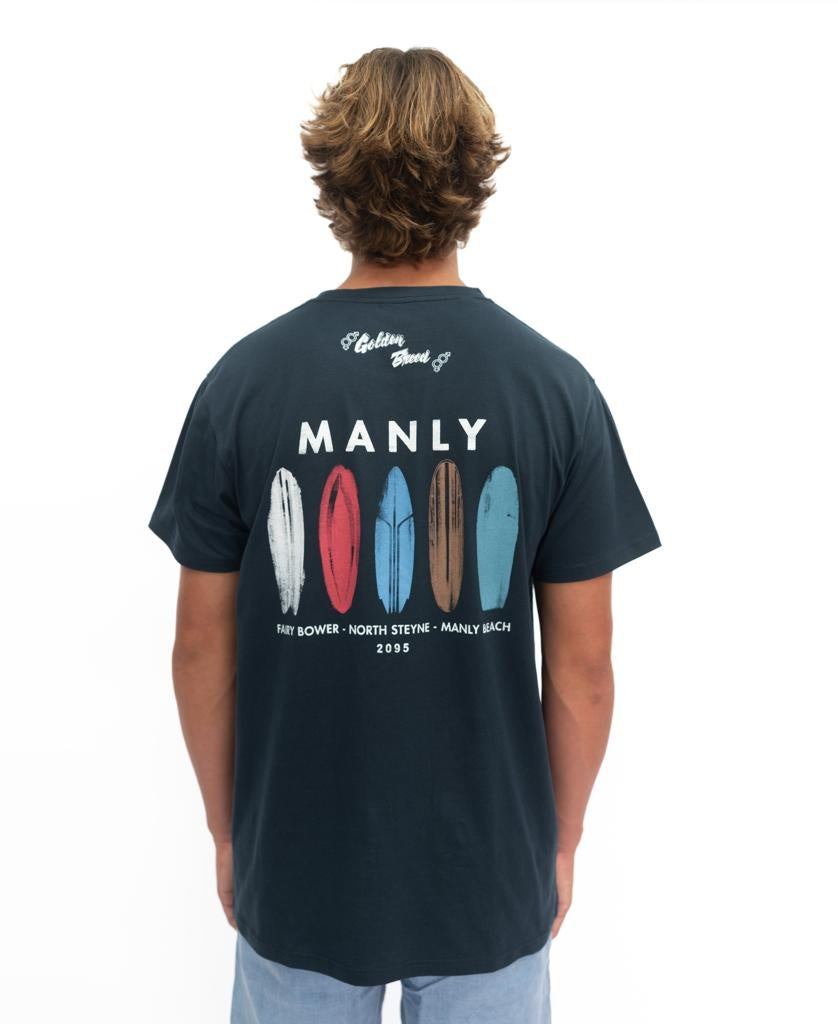 A person wears the Golden Breed Manly Boards Tee in navy—crafted from soft cotton with colorful surfboards and MANLY on the back—posing against a white background, perfect for laid-back surf town vibes.