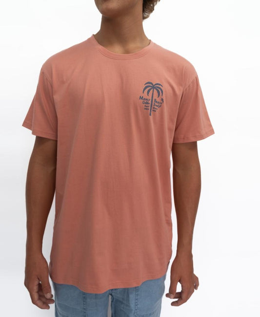 A person wearing the Golden Breed Manly Palm Loco Tee in Rose, a 100% cotton salmon T-shirt featuring a small palm tree and Miami Beach graphic on the left chest, pairs it with light blue jeans against a white background.