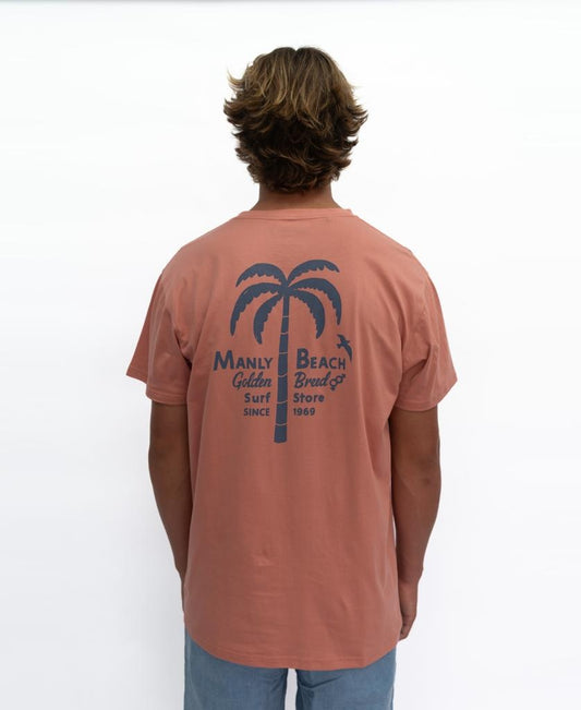 A person faces away, wearing the Manly Palm Loco Tee | Rose by Golden Breed—a 100% cotton pink tee featuring a blue palm tree graphic and Manly Beach Golden Breed Surf Store Since 1969 text.