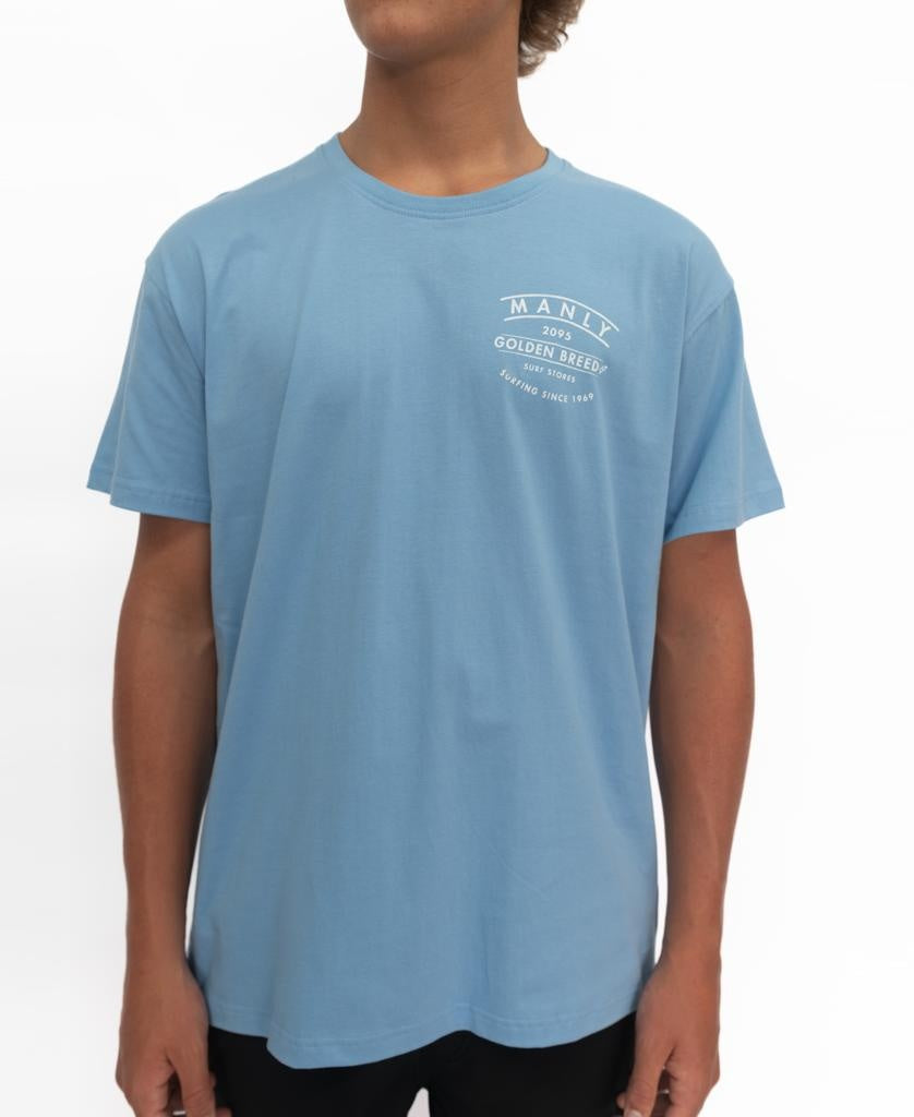 A person wears the Golden Breed Manly Boards Tee in Azure Blue, showing white printed text and a Surf Town logo on the left chest, standing against a plain white background.