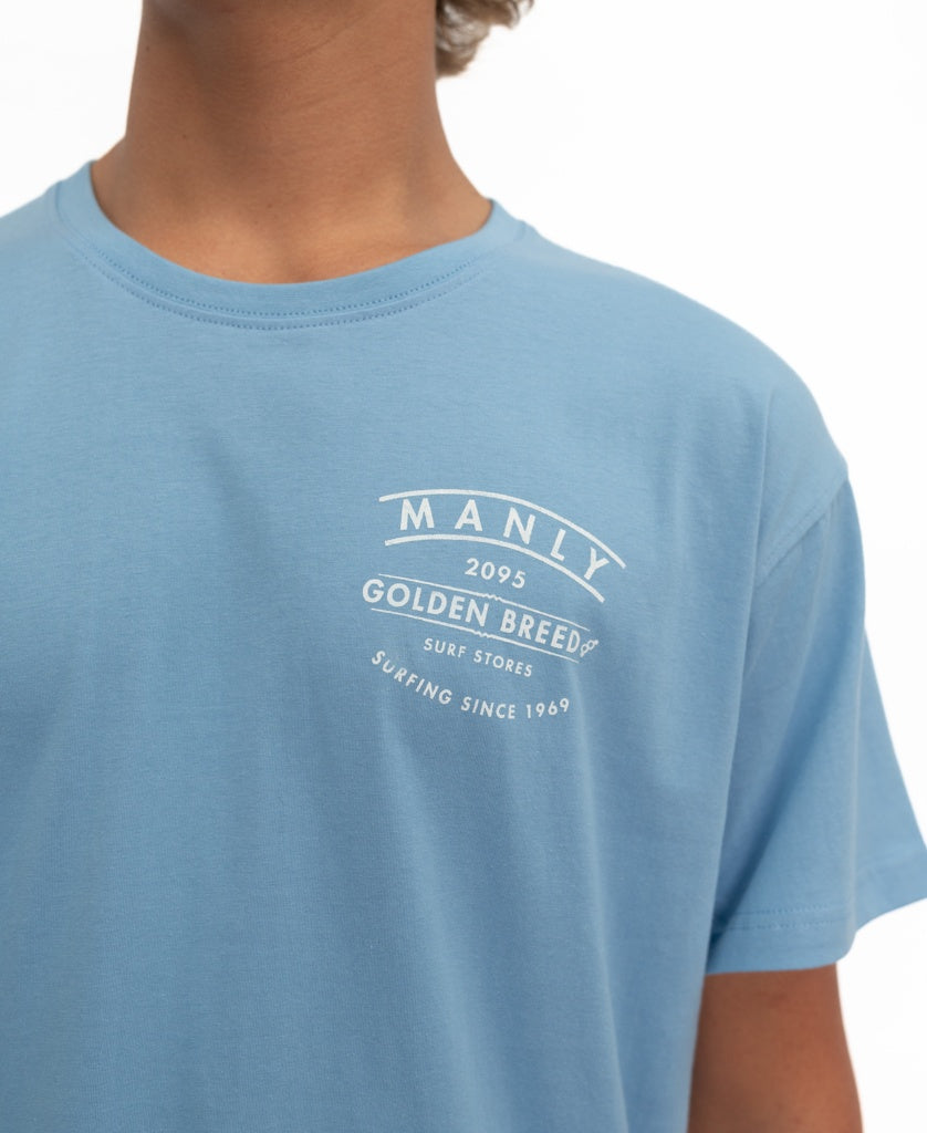 Wearing the Manly Boards Tee | Azure Blue by Golden Breed, featuring white MANLY 2095, Golden Breed Surf Stores, Surfing Since 1969 text on the left chest—a classic surf town style.