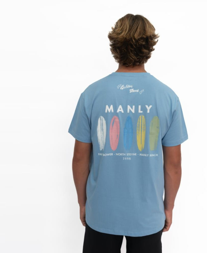 A person wearing the Golden Breed Manly Boards Tee in Azure Blue, which features five surfboards on the front and MANLY plus popular Surf Town beach locations printed on the back.