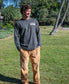 A man stands on grass wearing a long-sleeve dark shirt with text and Golden Breeds George Cord Pant in Dark Camel, featuring wide legs and a patch pocket, set against trees and a walkway.