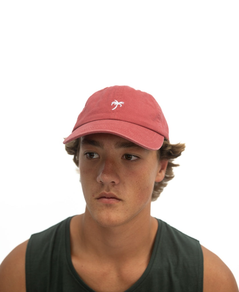 A young person wears the Golden Breed Palm Cap | Wash Red—a pink baseball cap with a small white palm tree logo—and a dark tank top, set against a plain white background.