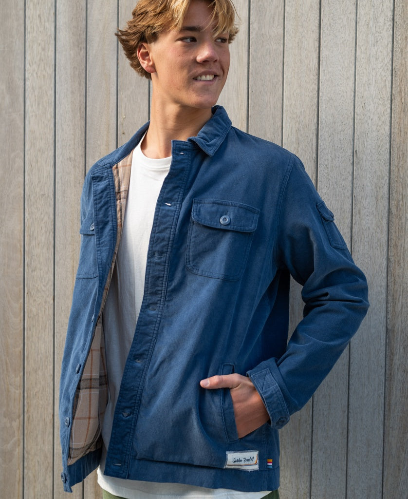 A young man wearing the Golden Breed Cahoots Cord Jacket in Vintage Navy over a white t-shirt stands before a wooden wall, gazing to the side with one hand in his pocket.