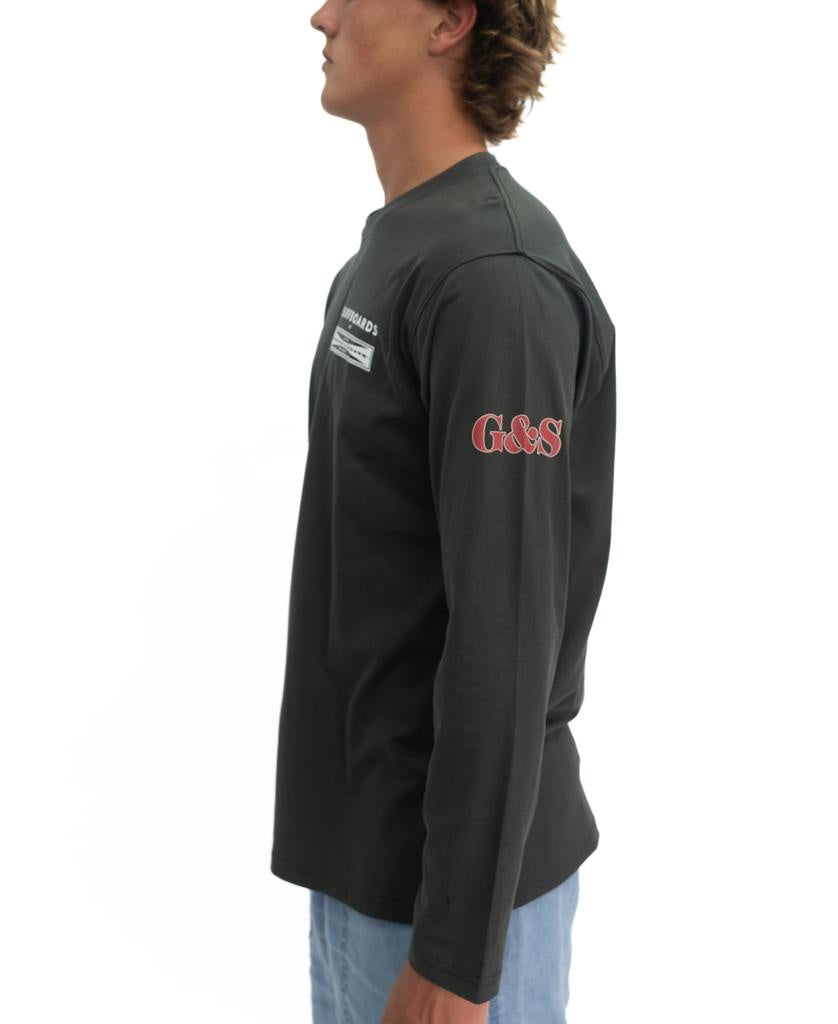 Man in a wash black G&S Surfboards Comp LS Tee by Gordon & Smith, with Go&S on the left sleeve, stands sideways against a white background—echoing surf heritage and the brand’s premium quality.
