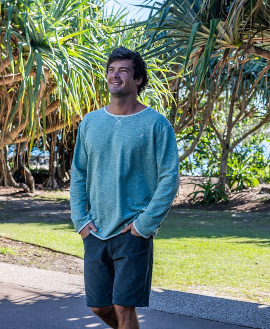 A man in a Surf Green Marle Classic Rag Top by Golden Breed and dark shorts strolls outdoors on a sunny day amid lush greenery, embodying an effortless surf style.