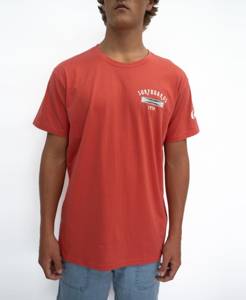 A person wearing a wash red G&S Surfboards Tee by Gordon & Smith featuring a small SURFBOARDS 1993 chest logo and light blue shorts, standing against a plain white background.