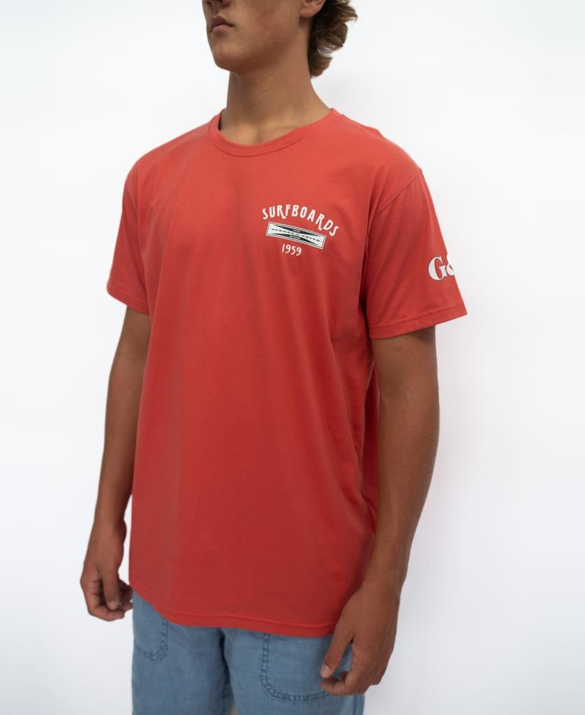 A person wears the Gordon & Smith G&S Surfboards Tee in Wash Red with light blue shorts, standing against a plain white background.