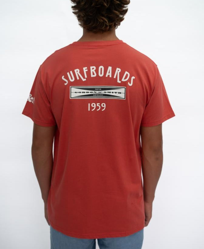 A person stands with their back to the camera, wearing a Gordon & Smith G&S Surfboards Tee in Wash Red with SURFBOARDS 1959 on the back, paired with light blue shorts against a plain white background.